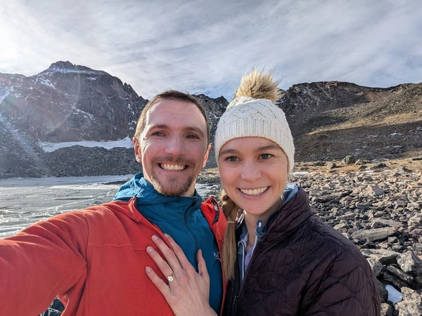 A smiling couple in winter gear posing by a frozen alpine lake with snow-capped mountains in the background