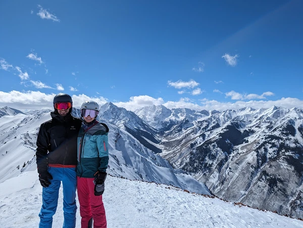 Two skiers in winter gear standing on snowy mountain peak with snow-covered peaks in background