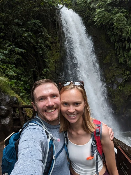 Couple smiling at camera with a tall waterfall and lush green vegetation in background