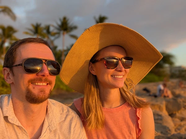 A smiling couple wearing sunglasses at sunset on a tropical beach with palm trees in the background