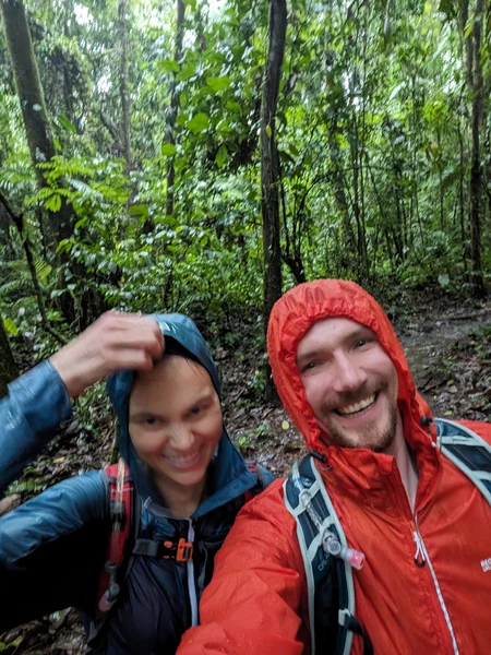 Two hikers smiling at camera in rainy forest wearing rain jackets and backpacks