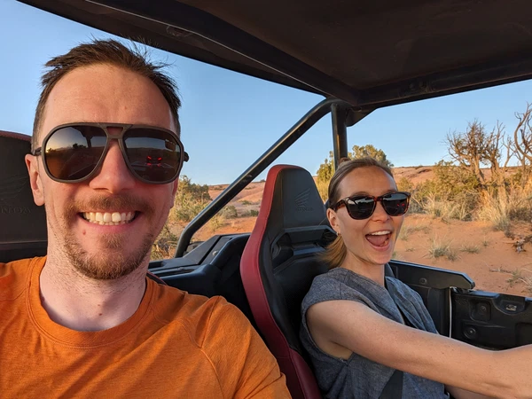 Two smiling people wearing sunglasses in a convertible vehicle in a desert landscape